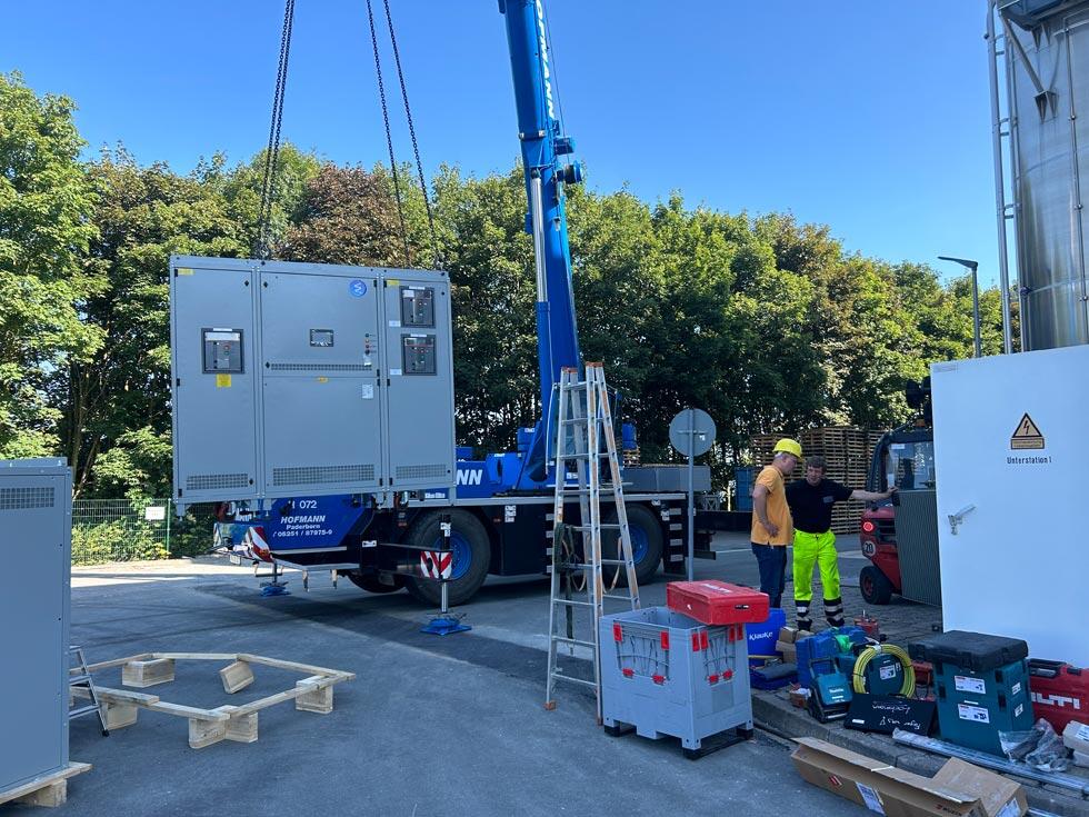Installation der Aussenstation - ein Teil der Installation hängt an einem Kran und 2 Bauarbeiter stehen diskutierend daneben.