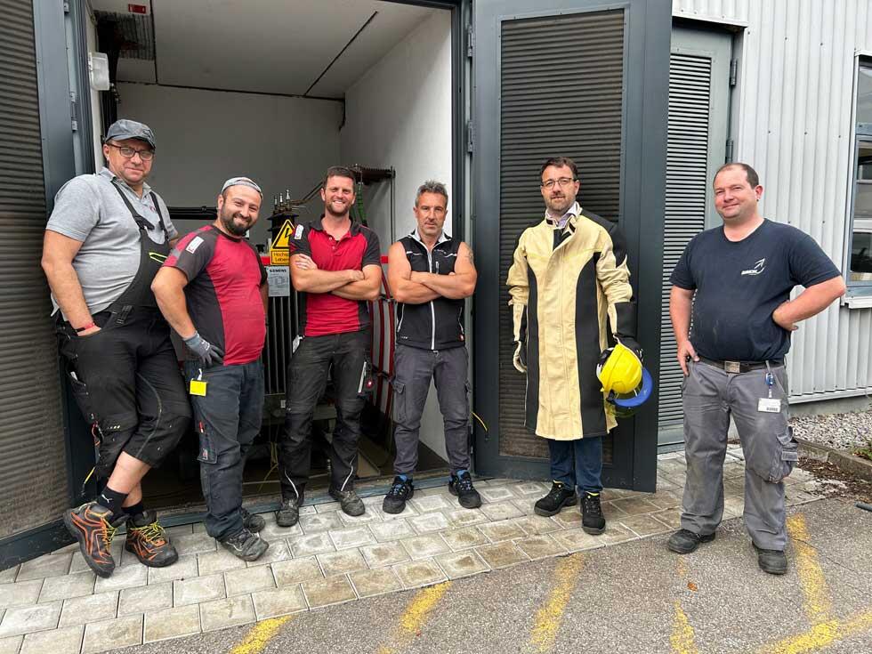 Sechs Installationsmonteure stehen vor einem Gebäude mit geöffneten Toren