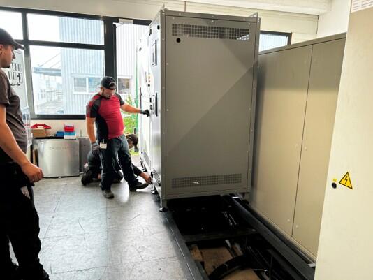 Transformator in einem Raum, eine Person steht und eine Person kniet davor, eine weitere Person sthet links und beobachtet