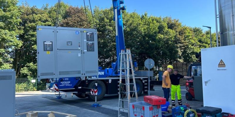 Installation der Aussenstation - ein Teil der Installation hängt an einem Kran und 2 Bauarbeiter stehen diskutierend daneben.