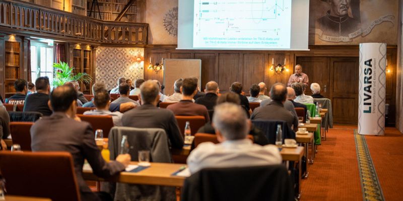 Personen sitzen auf Stühlen an Tischen, hintereinander aufgereiht und hören einem Vortrag zu. Vorne ist eine große Leinwand mit einer Abbildung.