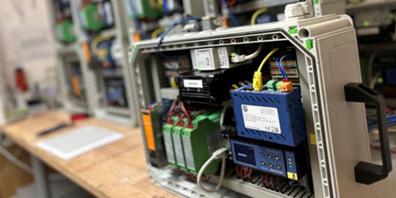 Grauer Kasten mit Kabeln und Anschlüssen auf einem Tisch, im Hintergrund weitere aufgestapelte Kästen.